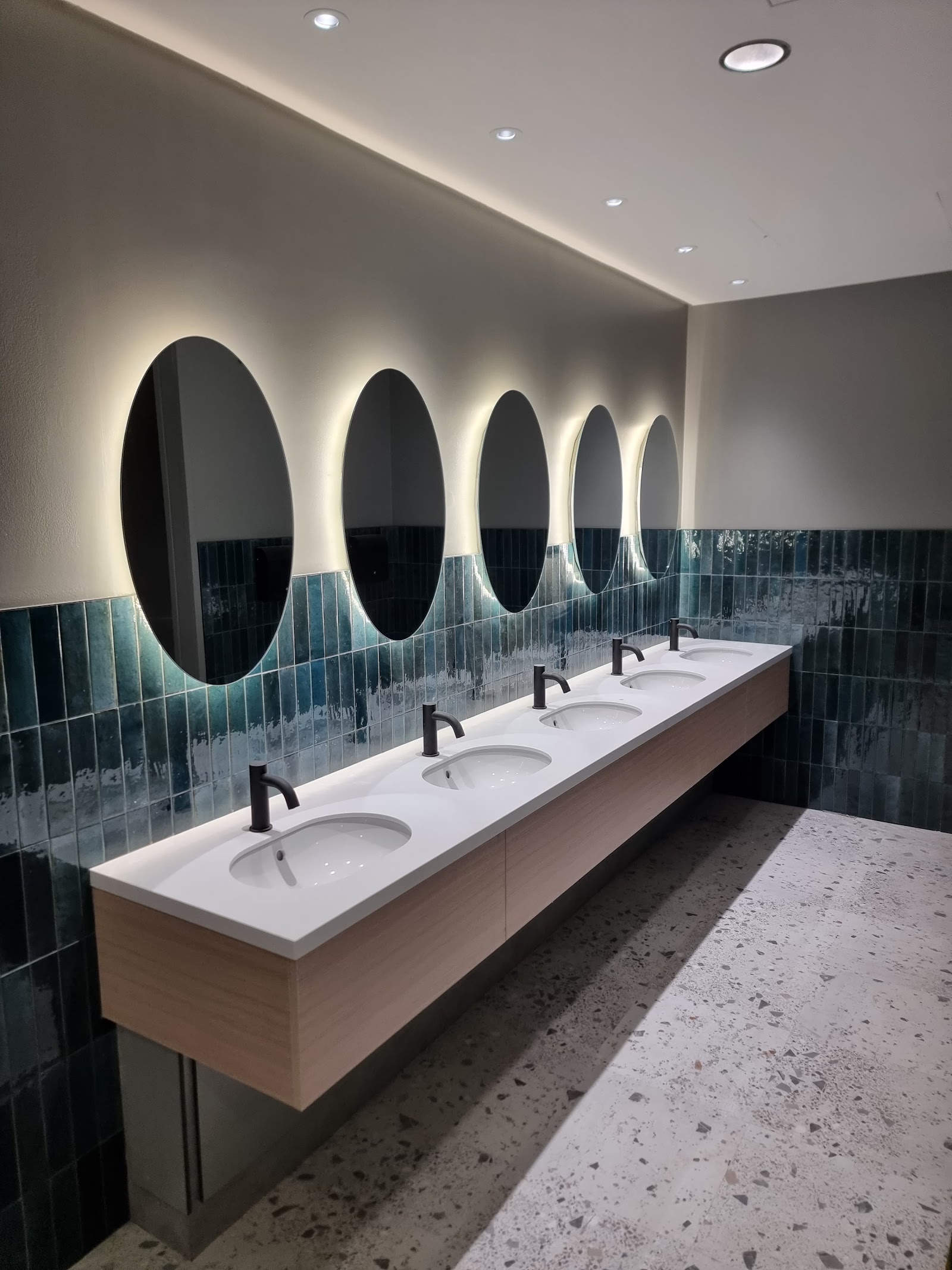A commercial washroom fit-out — a long trough basin with concealed taps beneath a run of backlit mirrors and glazed teal tiling.